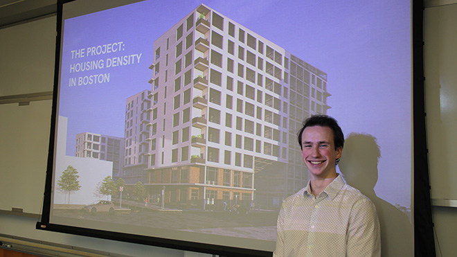 Senior Ryan Spillane stands, smiling, in front of a screen displaying the title slide of his housing density project