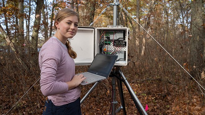 Sophomore Mandi Greenhalgh holding a laptop in front of a data logger box in the woods in Warren, Rhode Island