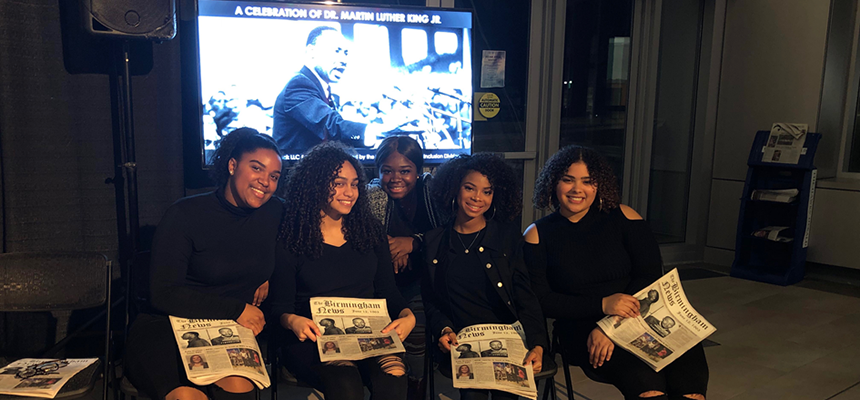 image of 5 students sitting in a row, holding a newspaper with a picture of Martin Luther King Jr. for a Black History Month performance.
