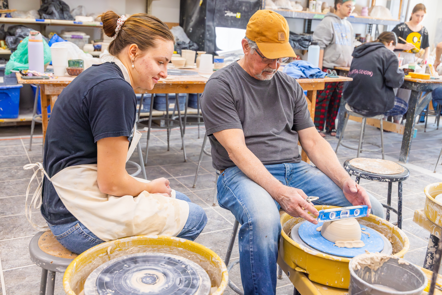 Leonti works with a student at the wheel, using a level atop a mound of clay to demonstrate precision, preparation, and technique.