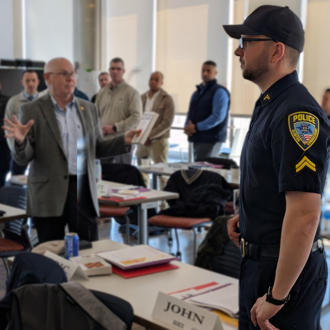 A police officer stands in a classroom while a man speaks to the group