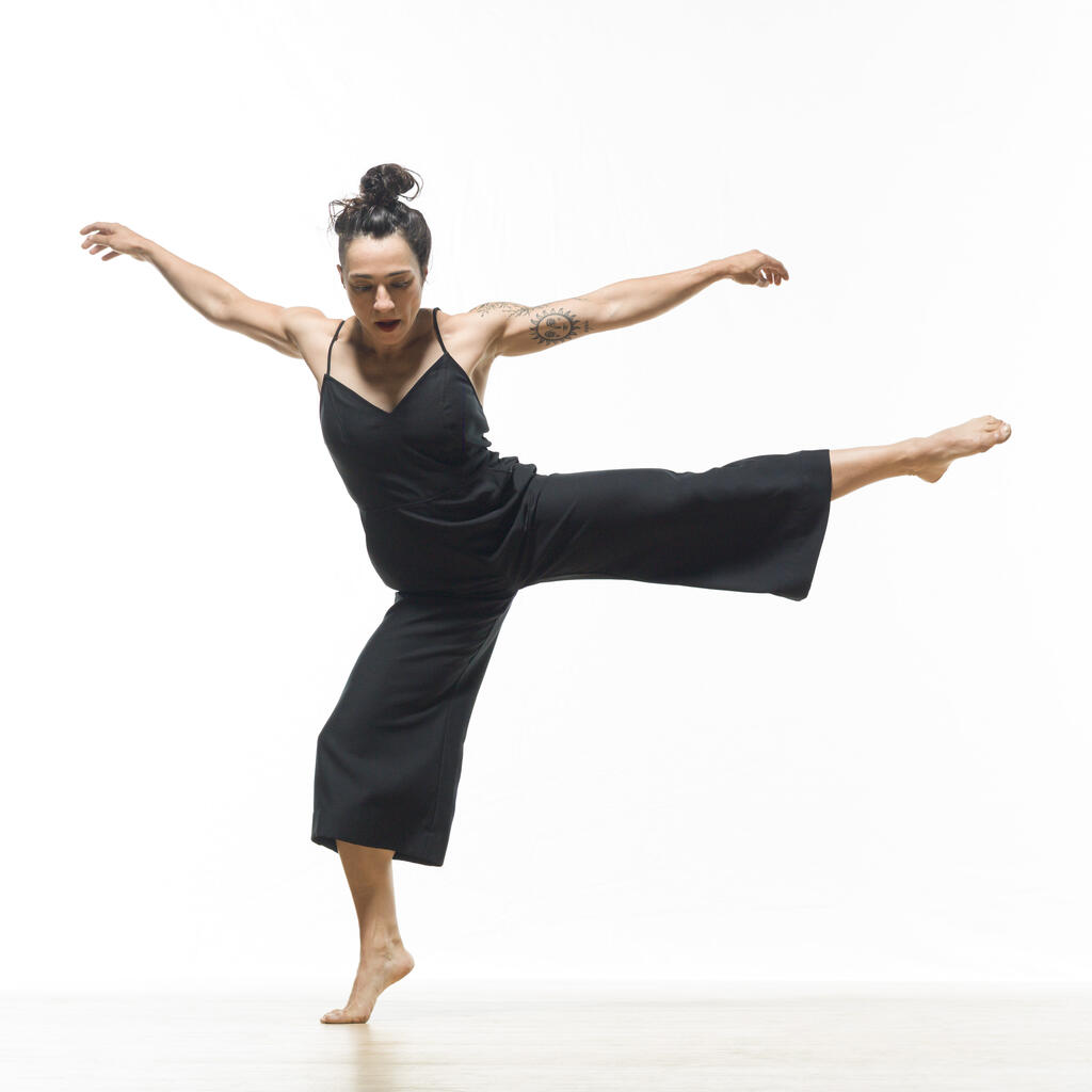 A dancer in a black outfit balances on one foot with arms extended and one leg raised against a white background