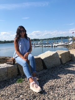 Student that identifies as female, sitting on rocks near teh ocean during teh day, wearing sunglasses and a blouse with jeans and sneakers. 