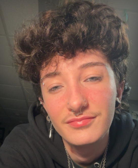 Person with short curly dark hair, facial piercings, and a black hoodie