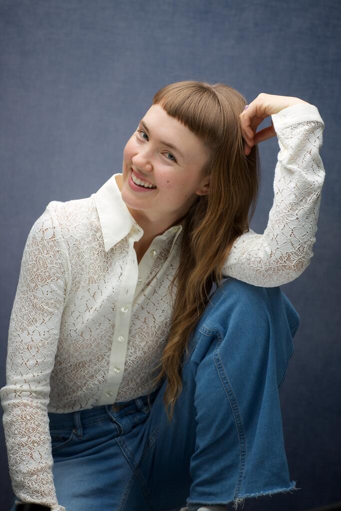 A smiling woman with long brown hair sits against a blue background, wearing a white lace shirt and blue jeans
