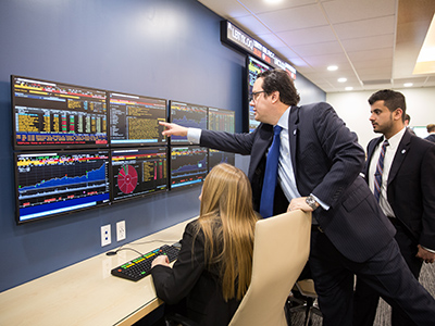 A professor pointing to a screen in the trading room as two students look on