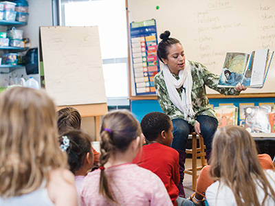 A student teacher sitting down and reading a picture book to a group of young students in a classroom