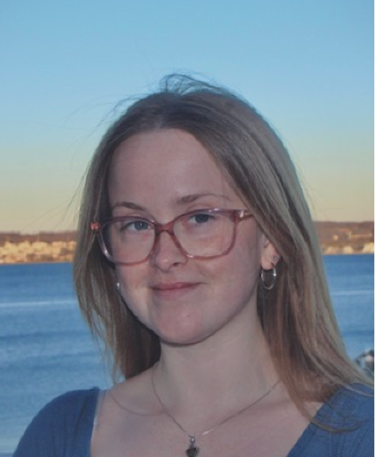 Person with long light brown hair and glasses standing in front of a blue waterfront