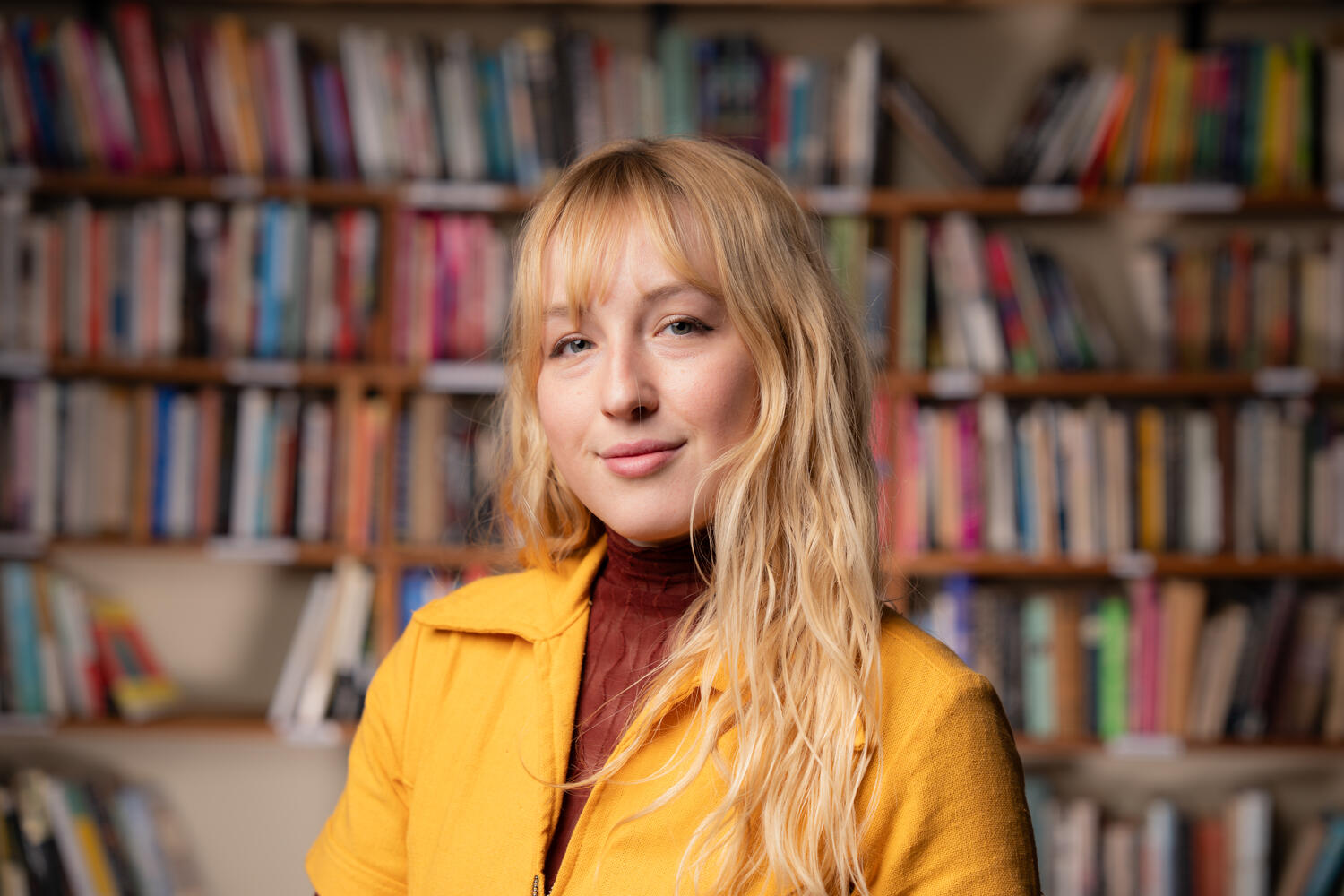A blonde woman in a yellow jacket poses in front of blurred bookshelves