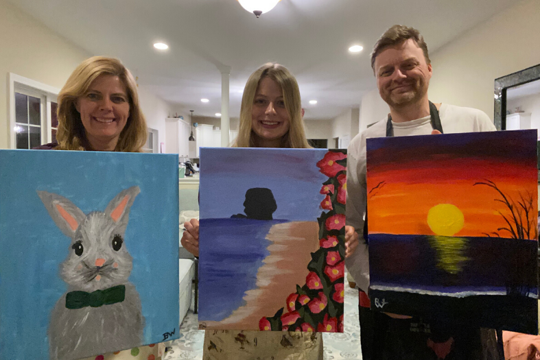 Three people pose, holding up paintings. 