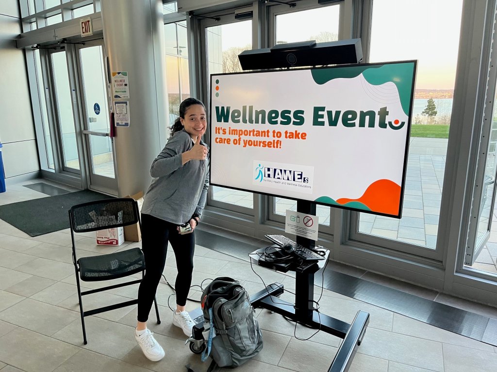Student smiling with thumbs up while standing next to giant TV screen that says Wellness Event