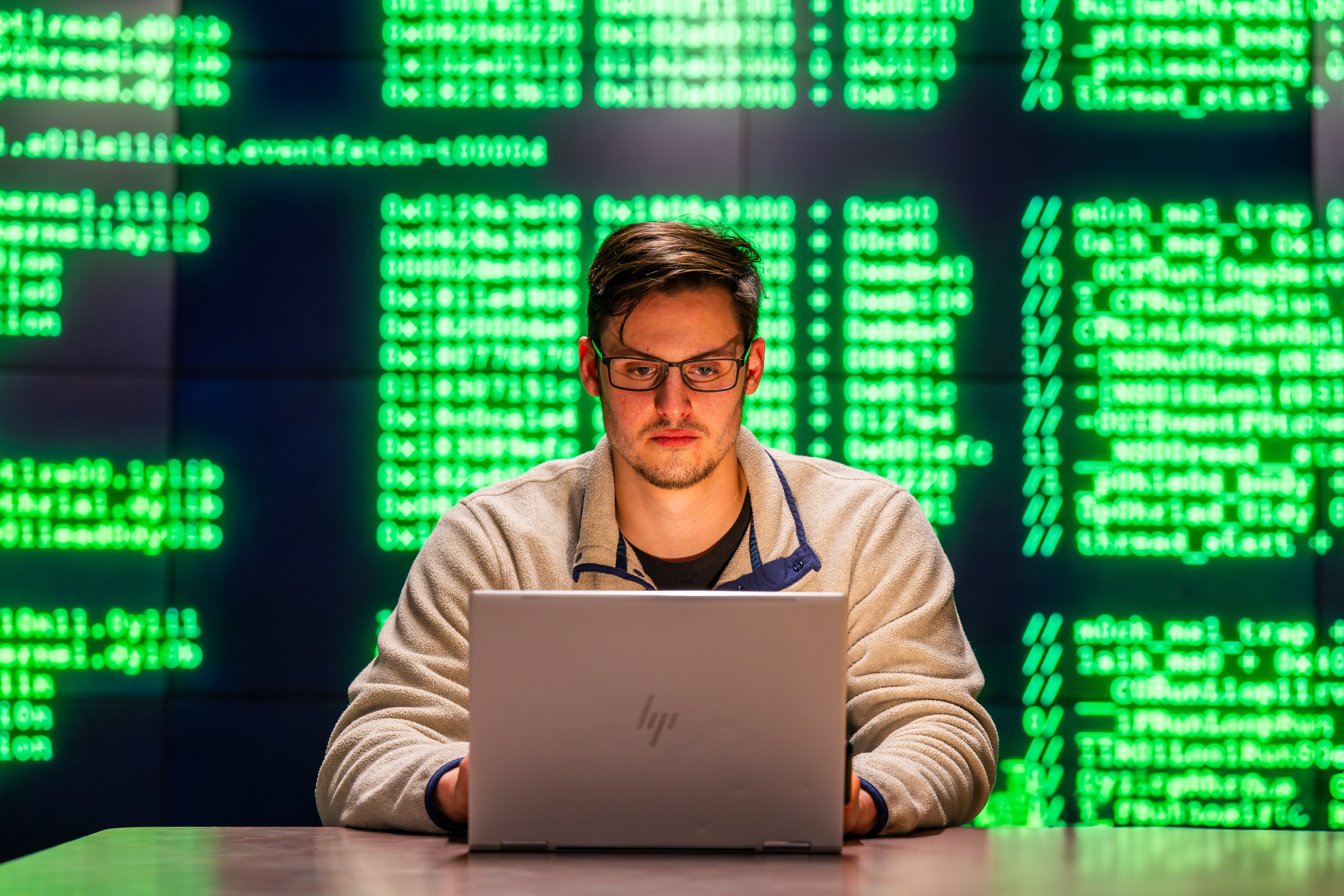 Student sitting at desk, in front of a background of various code, looking at laptop