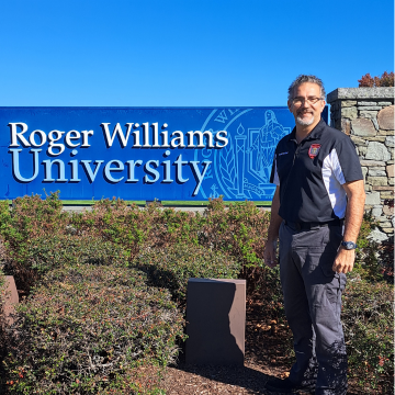 Alumni John Scott standing in front of the Roger Williams University sign outside on a day with a blue clear sky.