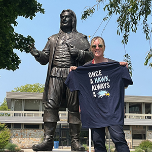 Kelly Scafariello holds a "Once a Hawk, always a Hawk" T-shirt by the Roger Williams statue on Bristol Campus