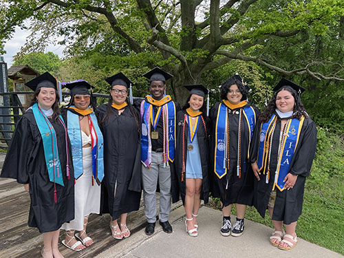 2025 RISE Scholars standing in front of a green tree wearing their graduation caps and gowns