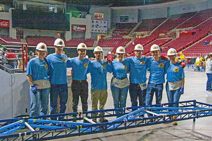 RWU Student Chapter of ASCE Steel Bridge Competition standing behind their steel bridge wearing blue t-shirts and white hard hats in the competition arena
