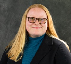 Allison Rivard smiling in a black sports coat and blue turtleneck sweater in front of a gray background.