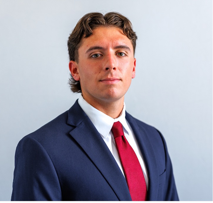 Rowan Conquest wearing a blue sports coat, white collared shirt, and red tie in front of a gray background.