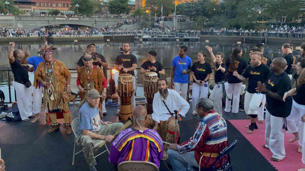 Foreground: Four members of Pomham Singers, some in traditional Eastern Woodlands garments, playing in a drum circle around a single low drum obscured from view. Middle Ground: On the left, two dancers with Pomham Singers in traditional Eastern Woodlands ceremonial garb. Behind them, three capoeira dancers with New Wave Martial Arts and Fitness move with the music. To their right, more members of New Wave Martial Arts and Fitness: two drummers playing djembes, then another large contingency of capoeira dancers moving to the music, all barefoot in white martial arts pants and black t-shirts. Background: Crowds sitting across the water from the performers, lining the waterfront at Waterplace Park.