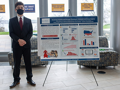 A student wearing a suit standing next to a poster presentation 