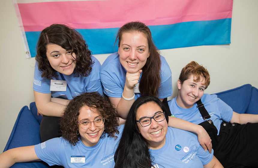 image of Current RWU students and staff in the Queer and Trans Resource and Advocacy Center in front of the trans pride flag