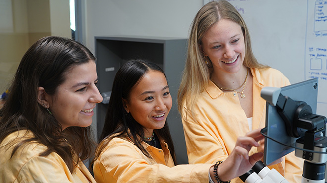 Three students in orange lab coats looking at an iPad attached to a microscope