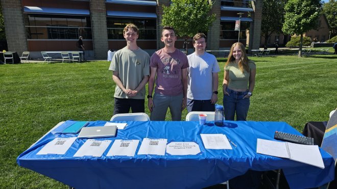 Four students stand behind their club table outside in the quad at the club fair.