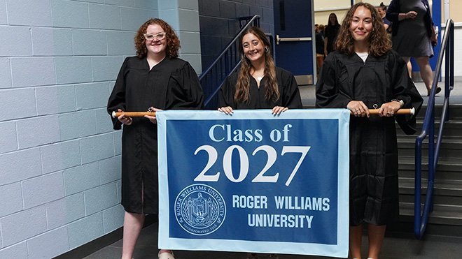 Three smiling students hold a blue "Class of 2027" flag in the RWU Recreation Center