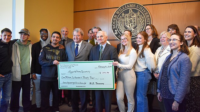 A photo of Senator Jack Reed holding a big check in the SECCM lobby, surrounded by administrators, faculty, and students