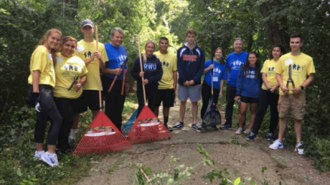 Group photo of students and president holding rakes on a path. 