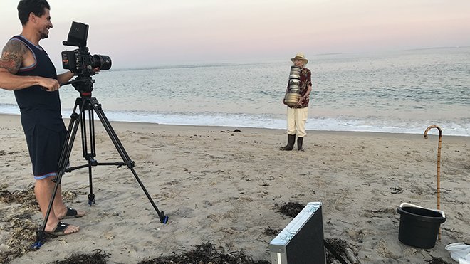 A director with camera films an actor holding film reels on a beach