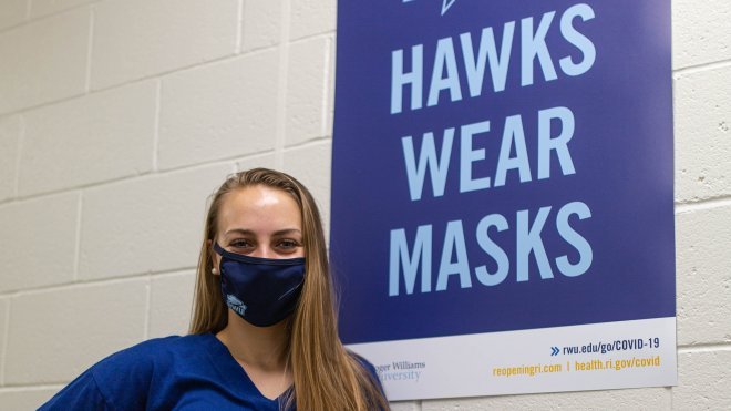 image of Jordan St.Onge, a student member of the Reopening Roger team, with a Hawks Wear Masks sign on campus