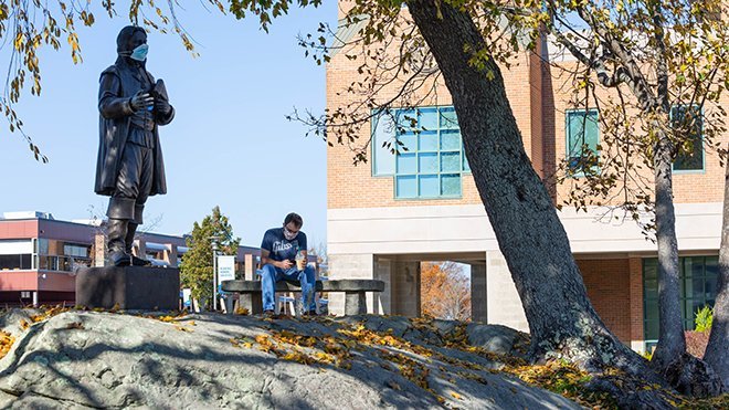 image of student sitting near Roger statue, both wearing masks