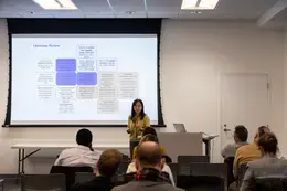 Presenter, Dr. Kara Li Liu, standing in front of project screen and talking in front of audience.