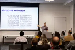 Presenter, Natalia Friedlander, standing behind desk with computer and gesturing towards the projector screen in front of an audience.