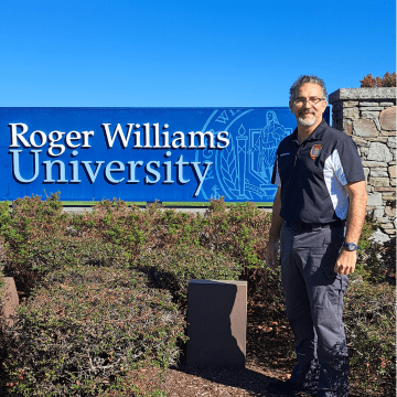 Alumni John Scott standing in front of the Roger Williams University sign outside on a day with a blue clear sky.