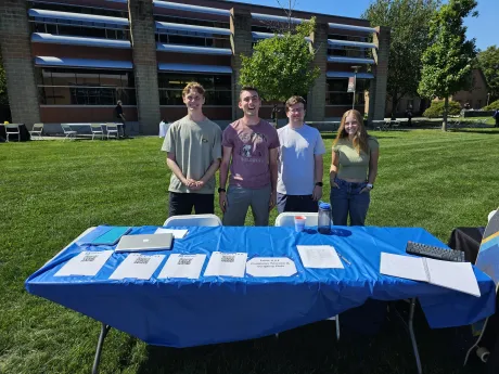 Four students stand behind their club table outside in the quad at the club fair.