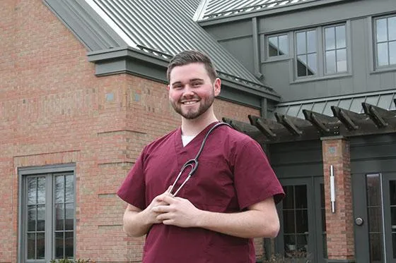 Student Cory Letendre stands in front of a building in medical scrubs.