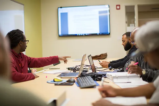 People sit in a classroom with laptops and a screen displaying information on the wall