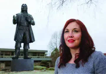 A woman with red hair stands outdoors beside a dark statue of a figure on a pedestal.
