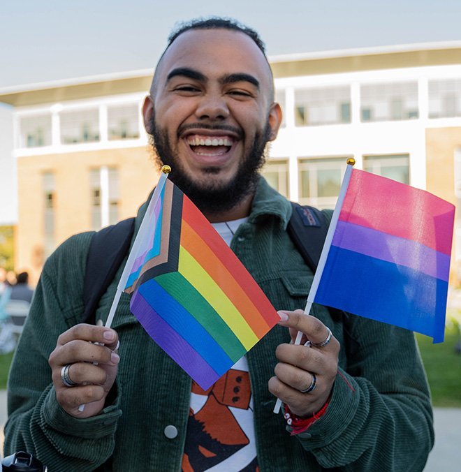 An RWU student smiles at the camera holding a Progress Pride flag and a Bisexual Pride flag