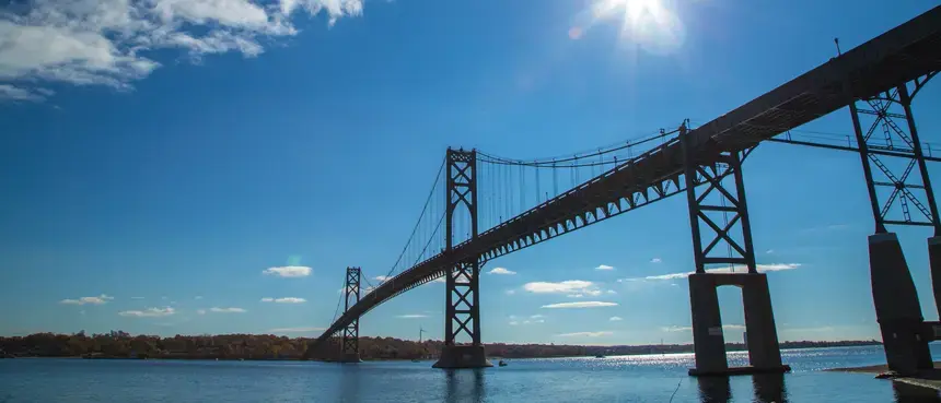 Photo of the Mt. Hope Bridge from a low angle, the sun is shining from the top right of the photo and reflecting off the ocean below the bridge.