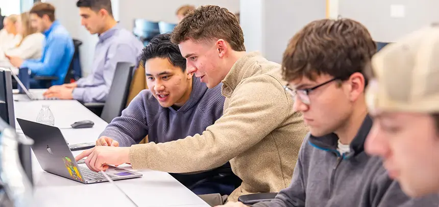 6 students pictured sitting at a table looking at laptops.