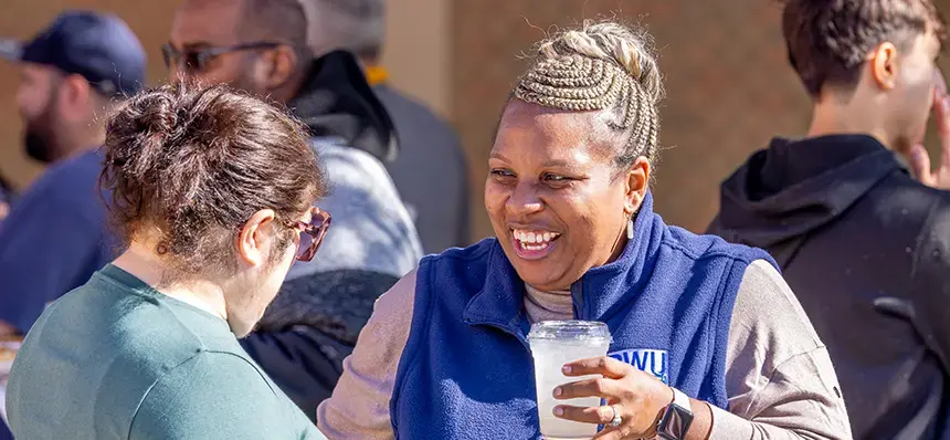 Two people talk to each other, one facing the camera. The one facing the camera is smiling and holding a beverage.