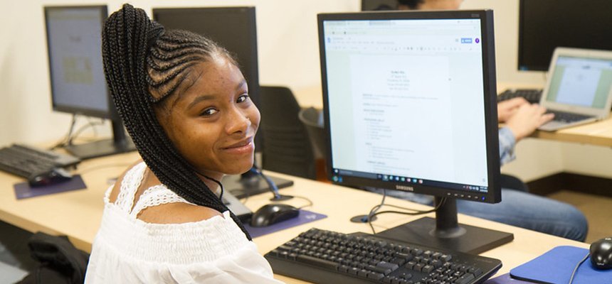 Student sits at computer and smiles at camera