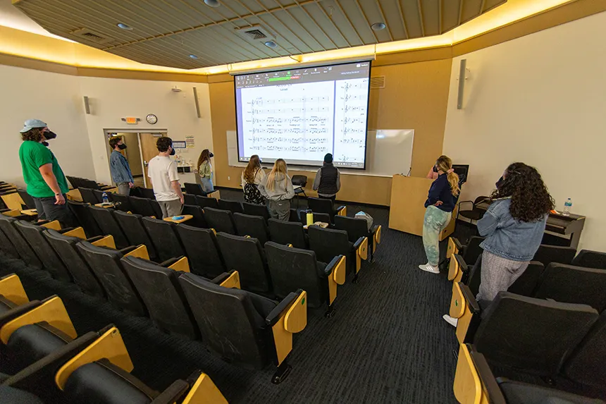 Image from back of hall of members of Hawkward a capella singing group at practice facing big screen
