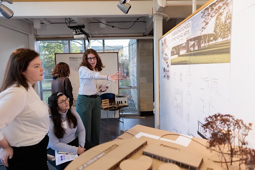 Three people stand in the Architecture gallery next to a 3D design of a building and a poster that shows the student's plans