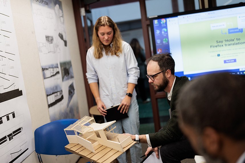A student stands next to a reviewer sitting down, both looking at a 3D model of a building