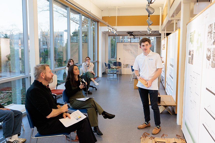 An Architecture student stands in the gallery next to his design plans as two reviewers sit in chairs and listen