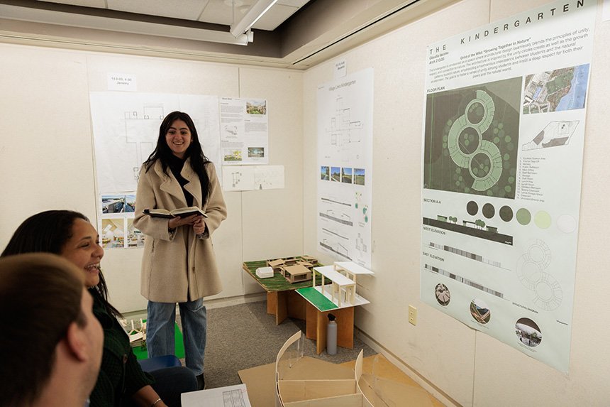 An Architecture student stands in a room next to her poster design as two reviewers sit